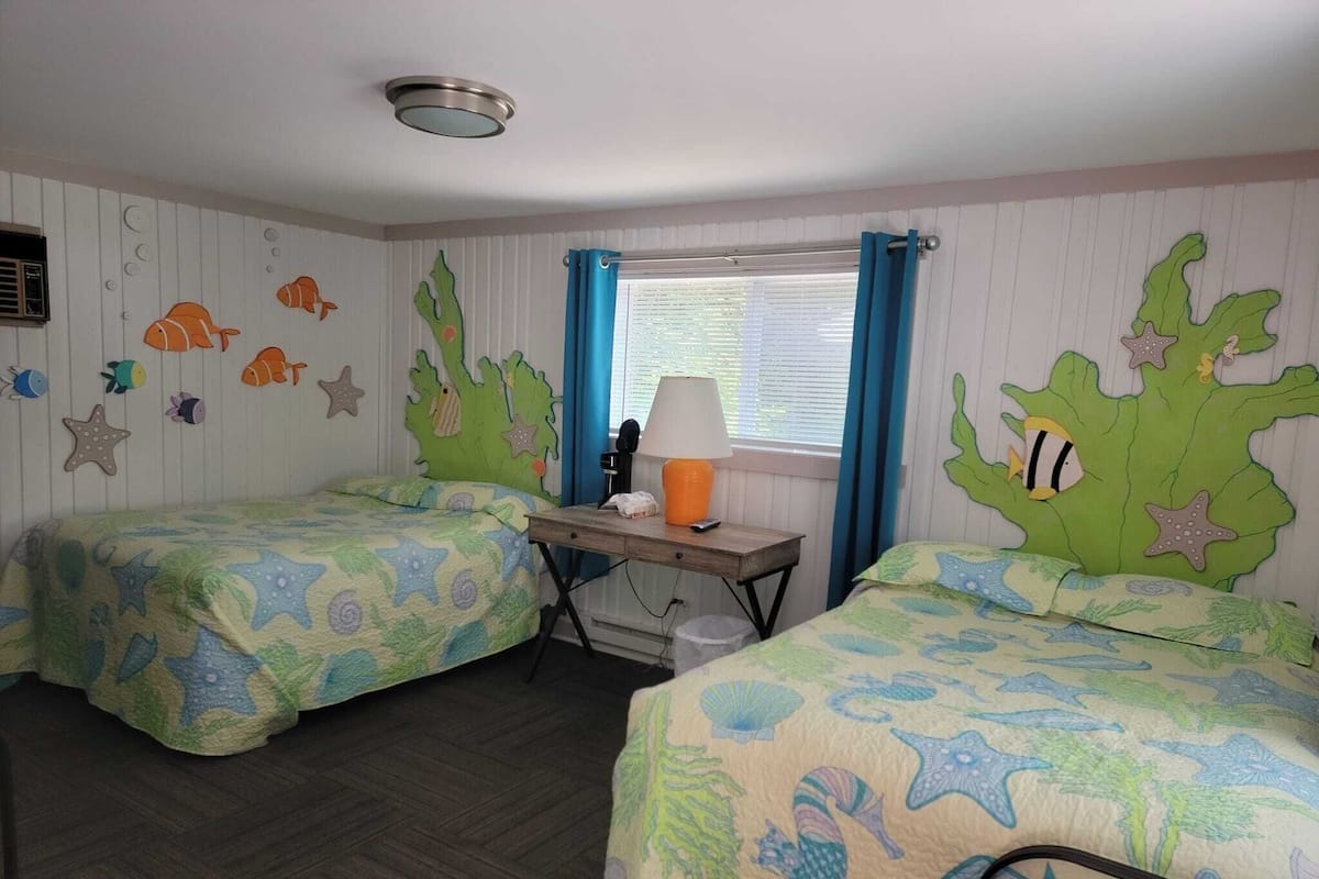 A room with two beds, each with a marine-themed bedspread, a desk with a lamp, and a window with blue curtains.