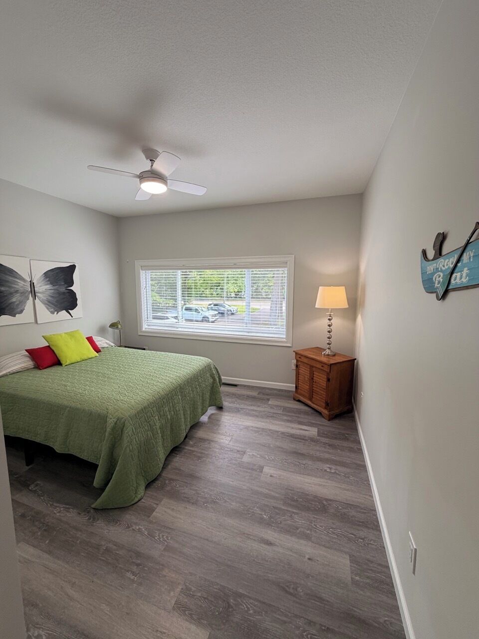 A bedroom with a bed, a ceiling fan, a window with blinds, a nightstand with a lamp, and artwork on the wall.