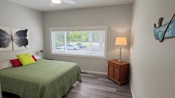 A bedroom with a bed, a ceiling fan, a window with blinds, a nightstand with a lamp, and artwork on the wall.