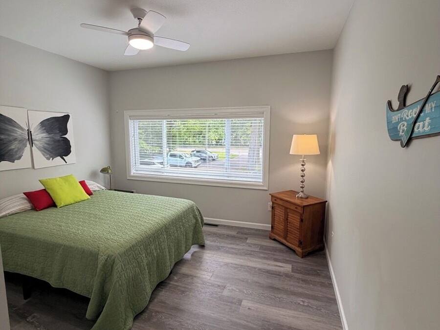 A bedroom with a bed, a ceiling fan, a window with blinds, a nightstand with a lamp, and artwork on the wall.