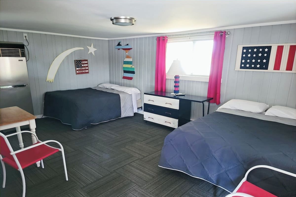 A room with two beds, a desk, a chair, a dresser, and a window with curtains.