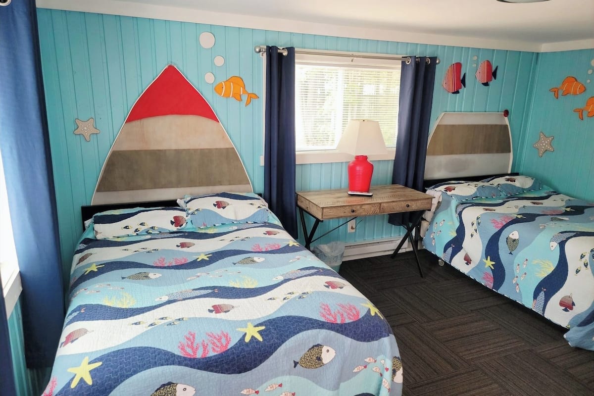 A room with two beds, each with a wave-patterned bedspread, a small table with a lamp, and a window with curtains.