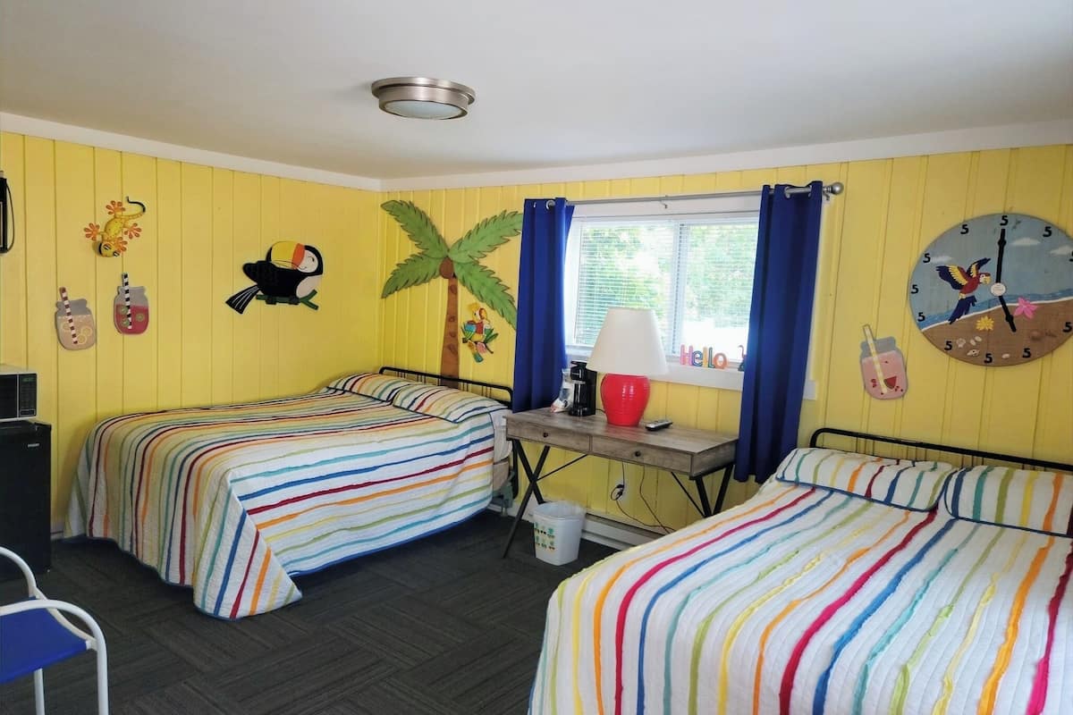 A room with two beds, a desk with a lamp, a window with curtains, and a wall clock.