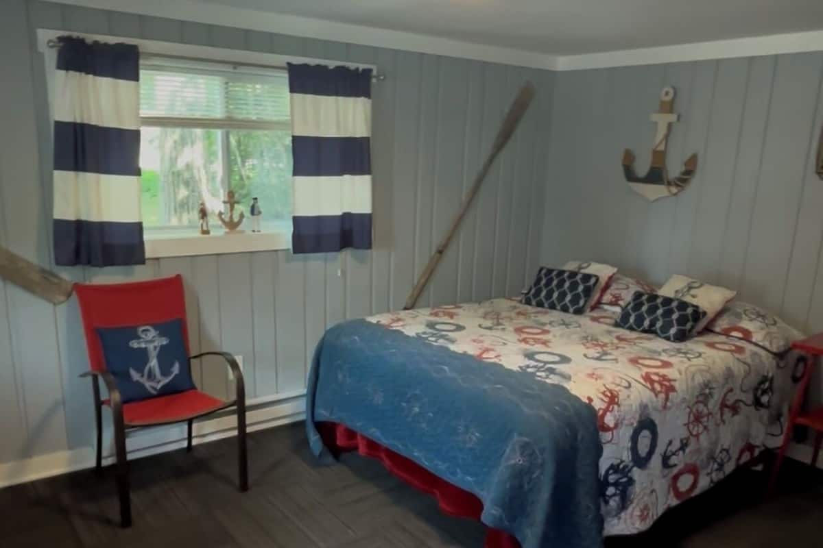 A bedroom with a bed, a chair, a window with curtains, a ceiling light, and a wall-mounted anchor decoration.