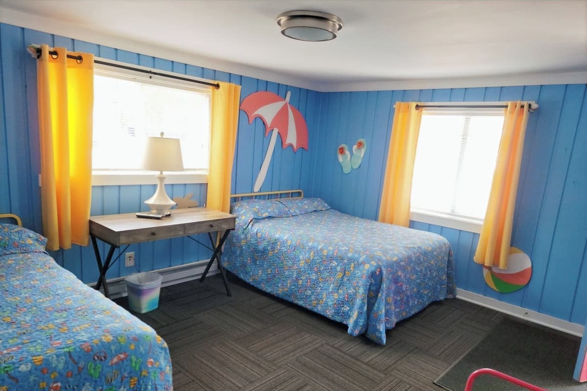 A room with two beds, a desk, and a chair. There are curtains on the windows, and a beach-themed wall decoration.