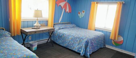 Una habitación con dos camas, un escritorio y una silla. Hay cortinas en las ventanas y una decoración de pared con temática de playa.