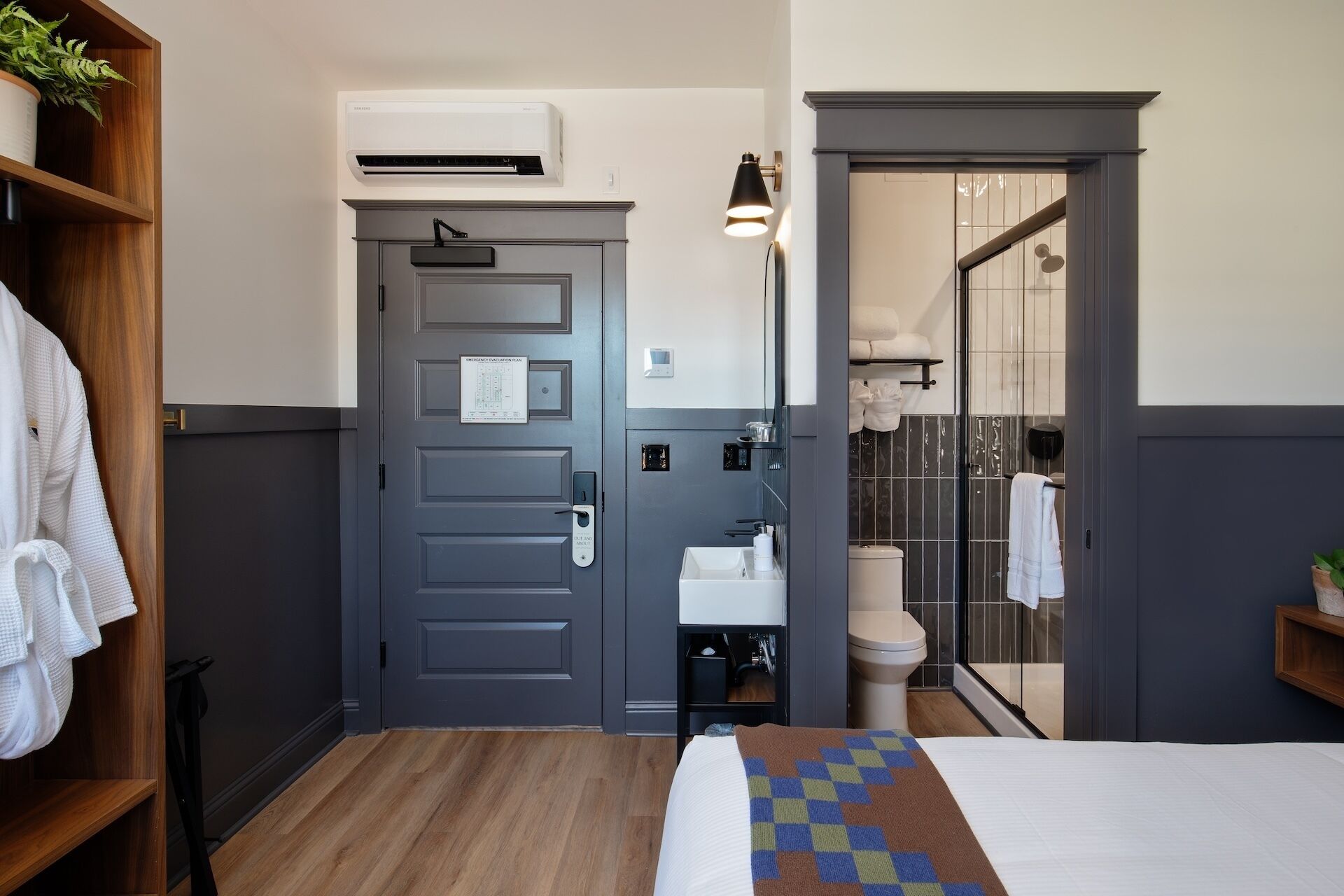 Habitación de hotel con una puerta gris, una mesita de noche con lámpara, una cama con manta estampada, un lavamanos pequeño blanco y una zona de ducha visible a través de una puerta de cristal.
