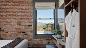A room with a brick wall, a window with a view of hills, a bed, a small stool, a wooden shelf with a laptop, and a towel rack. - Lightwell Hotel & Spa (Hood River)