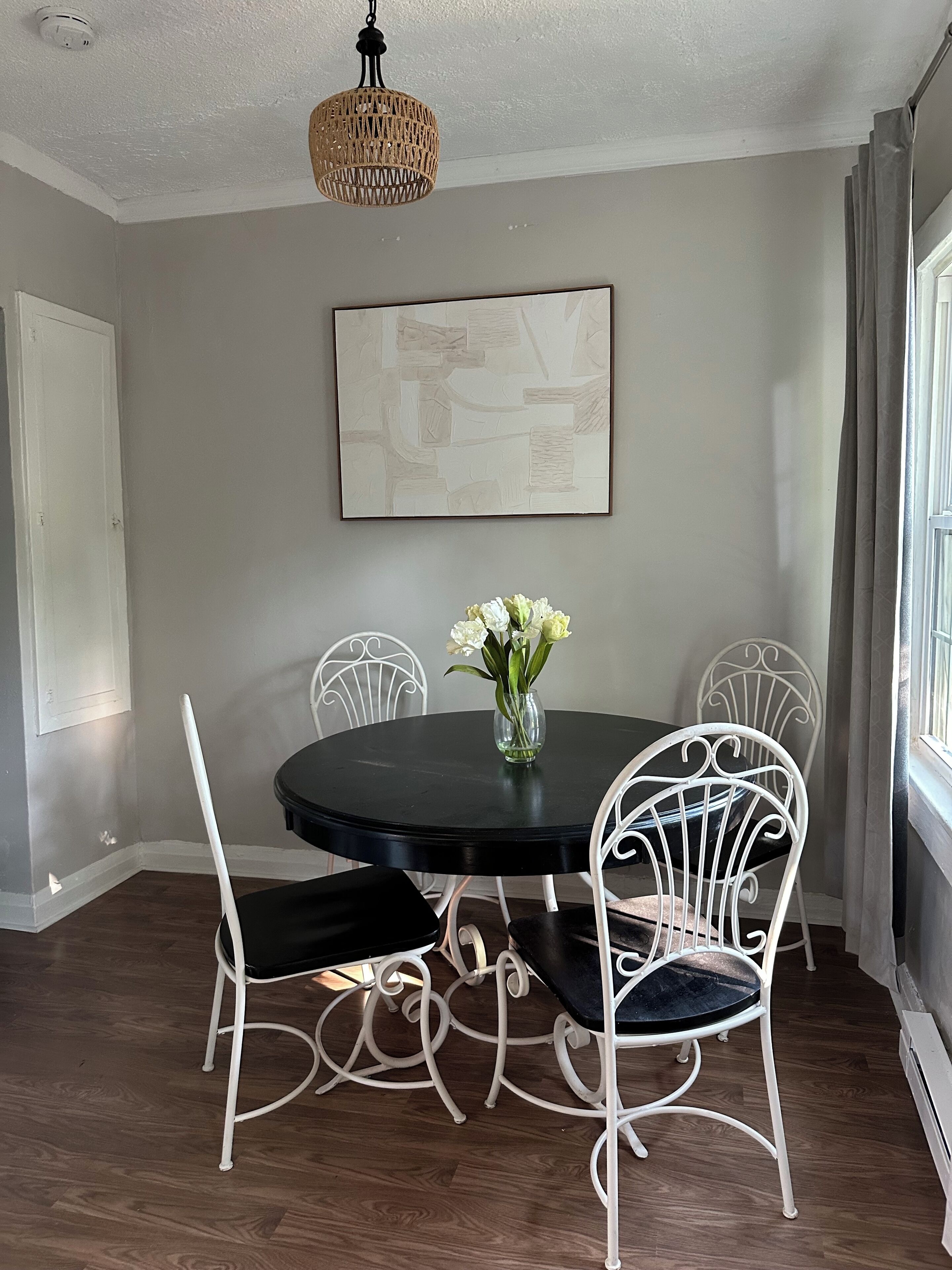 A dining area with a round table, white metal chairs, a vase with flowers, a hanging light fixture, and a framed abstract painting on the wall.