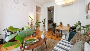 Een moderne woonkamer met een bank, salontafel en potplanten. Er is een eethoek met een tafel en stoelen, en een keuken op de achtergrond.