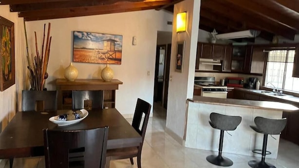 A dining area with a wooden table and chairs, a kitchen with a bar, and a painting on the wall.