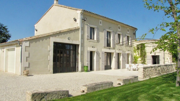 Un edificio de dos pisos con un amplio portón de garaje, un muro de piedra y una pequeña zona de patio con asientos exteriores.