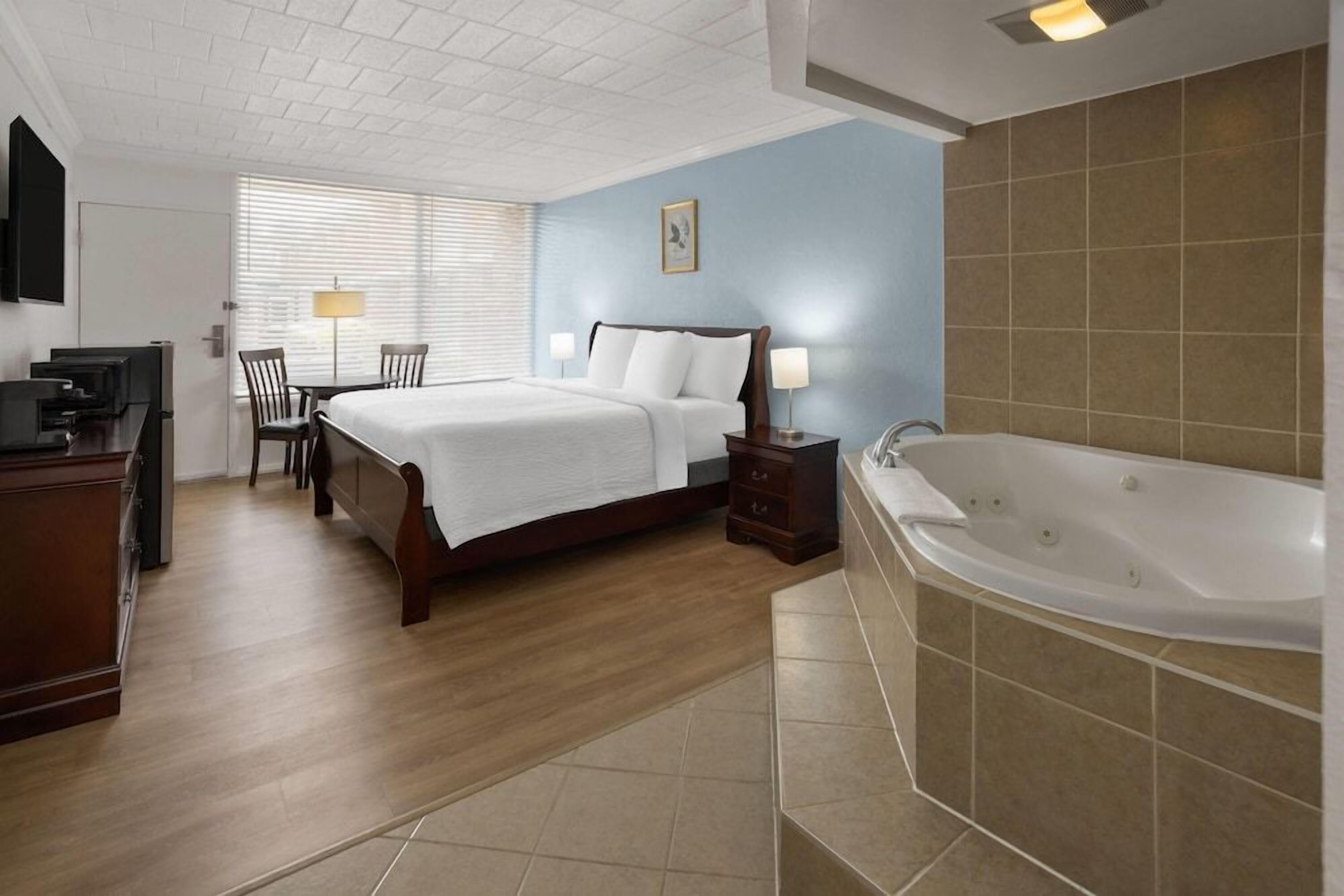 A hotel room with a bed, bedside table, lamp, and a large bathtub.