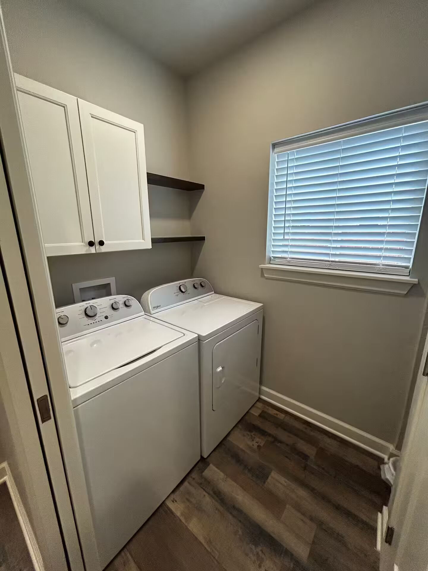 A laundry room with two washers and dryers, a window with blinds, and a built-in shelf above the dryers.