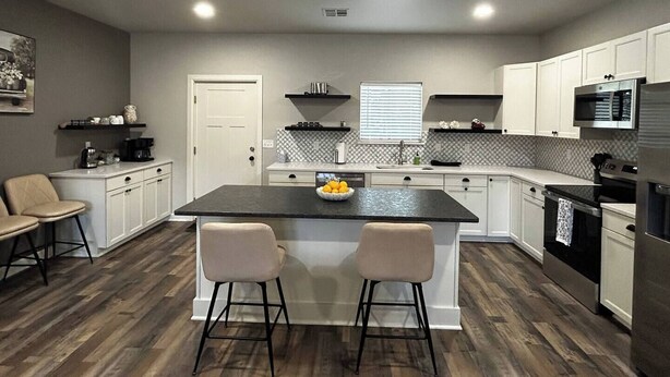 A modern kitchen with white cabinets, a black countertop island, two bar stools, and a window with blinds. - Silver Leaf (Stillwater)