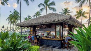 A thatched-roof bar with a stone-tiled floor, surrounded by lush greenery and palm trees.