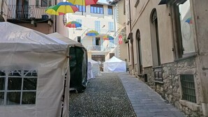 Uma rua estreita com calçada de pedras, edifícios com varandas e guarda-chuvas coloridos pendurados.