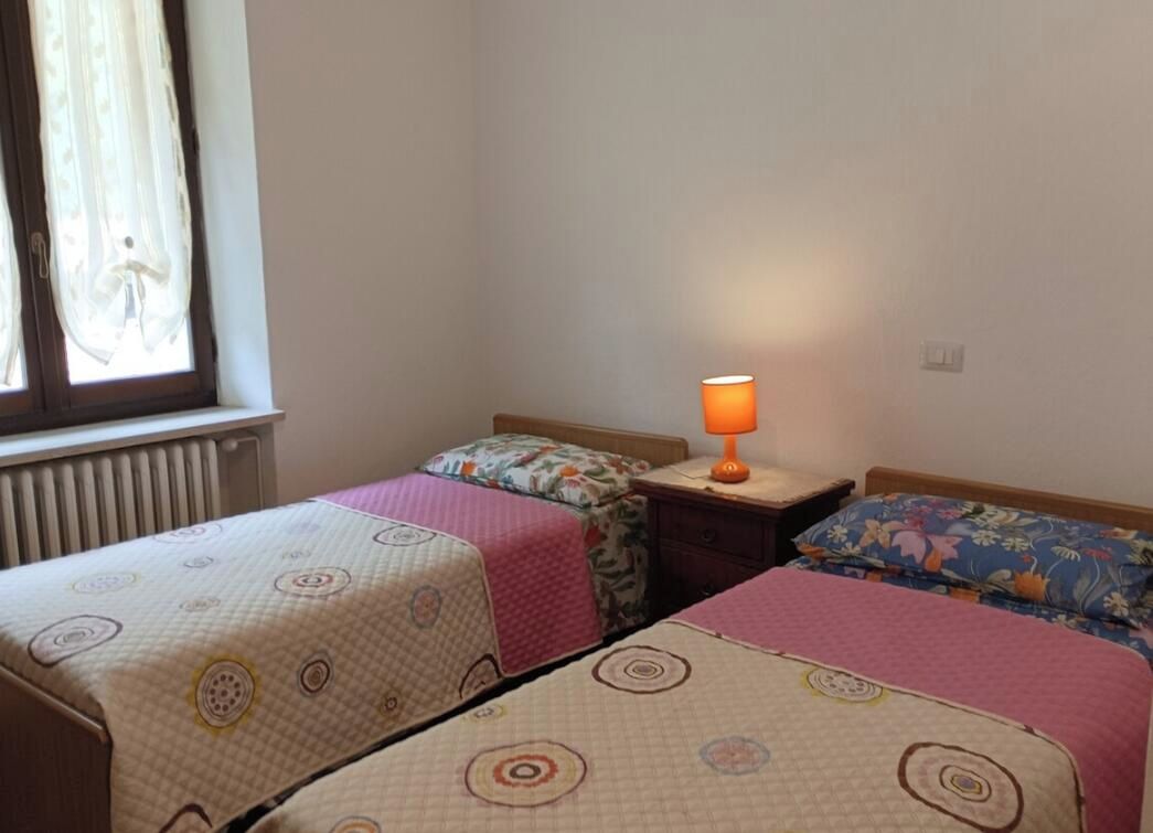 Ein Zimmer mit zwei Betten, jeweils mit einem gemusterten Bettbezug, einem Nachttisch mit Lampe und einem Fenster mit Vorhängen.