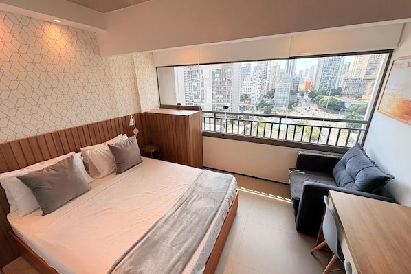 A modern bedroom with a large bed, a small sofa, a wooden desk, and a balcony with a city view. - On brooklin (Sao Paulo)