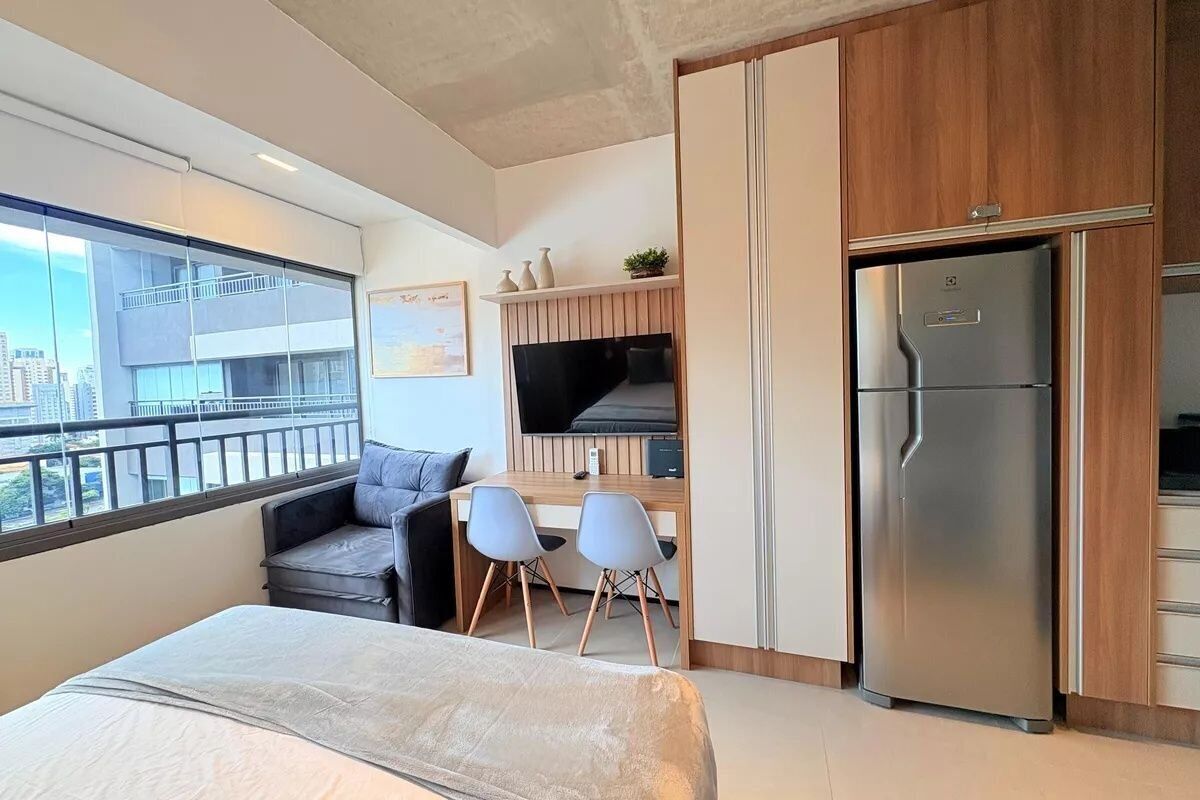 A modern hotel room with a bed, a desk with two chairs, a television, a refrigerator, and a balcony with a city view.