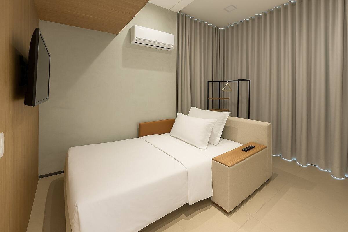 Ein Hotelzimmer mit einem Bett, einem an der Wand befestigten Fernseher, einer Klimaanlage und einem Vorhang mit beleuchtetem Saum.