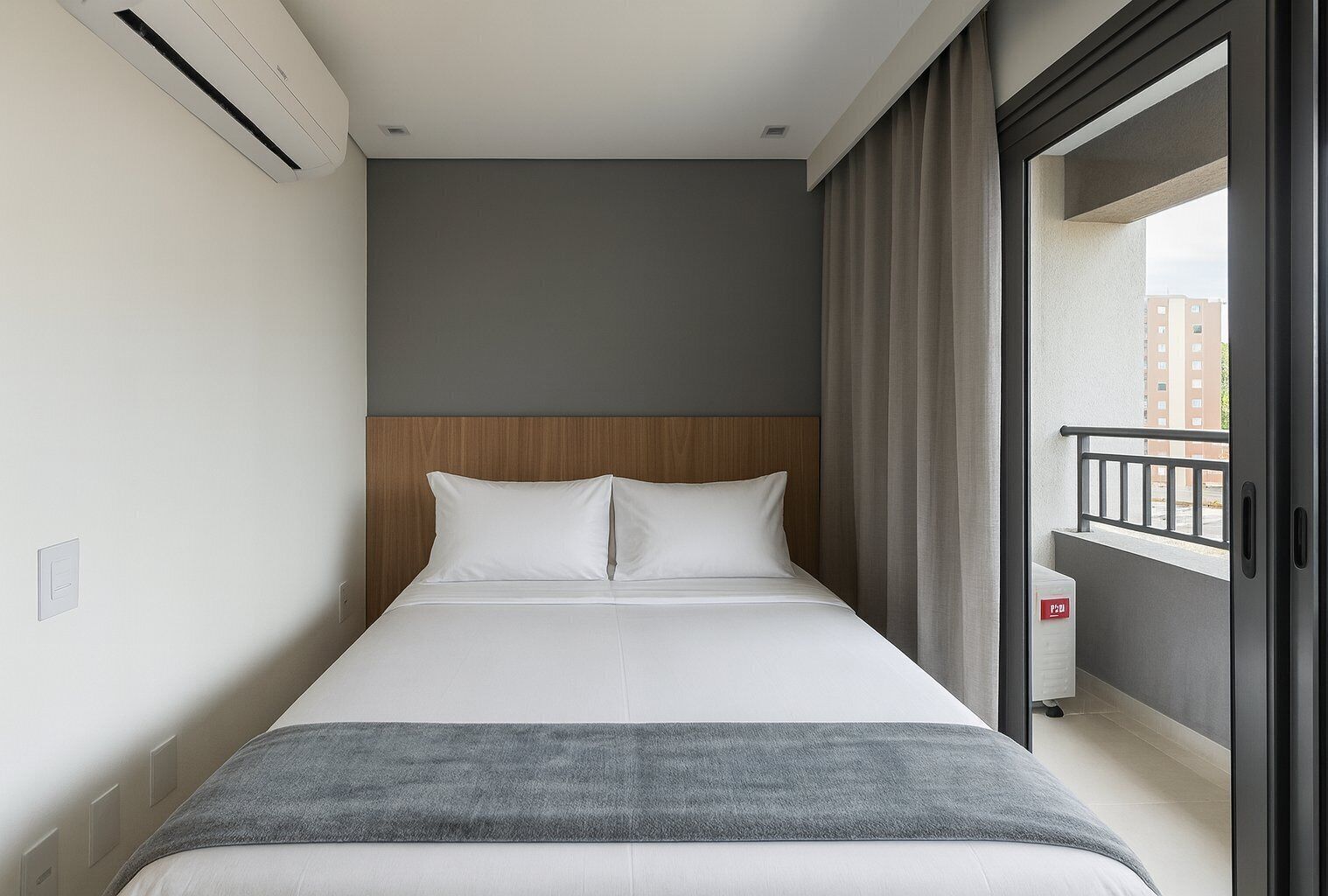 Une chambre d’hôtel avec un lit, une tête de lit grise, une couverture grise, une fenêtre avec des rideaux et une vue sur les bâtiments à l’extérieur.