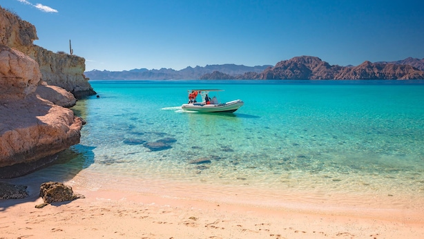 Un barco en un mar turquesa cristalino con acantilados rocosos y montañas al fondo.