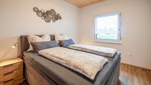 Um quarto com cama, mesas de cabeceira, abajur, janela e um objeto decorativo na parede.
