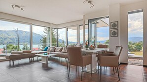 A spacious living room with a large window overlooking a lake and mountains, furnished with a sofa, chairs, and a coffee table.