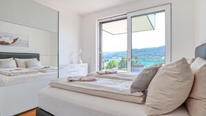 A modern bedroom with a large bed, a nightstand, a dresser, and a view of a lake and hills through a sliding glass door.