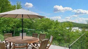 Eine Holzterrasse mit einem runden Tisch, Stühlen und einem Sonnenschirm, mit Blick auf einen See und einen Wald.
