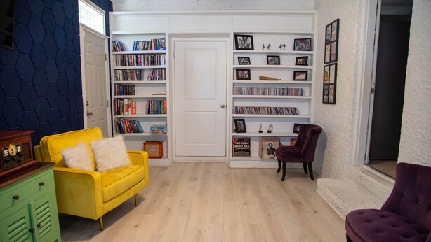 Une pièce avec un canapé jaune, un fauteuil violet et une bibliothèque remplie de livres et de DVD.