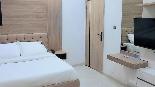 A modern bedroom with a tufted headboard, a flat-screen TV, and a wooden wardrobe. - Smart Home Hotel Lagos (Lekki)