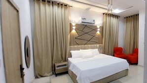 A modern hotel room with a large bed, a bedside table, a small nightstand, a wall-mounted air conditioner, and two red chairs. - Smart Home Hotel Lagos (Lekki)