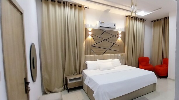 A modern hotel room with a large bed, a bedside table, a small nightstand, a wall-mounted air conditioner, and two red chairs. - Smart Home Hotel Lagos (Lekki)