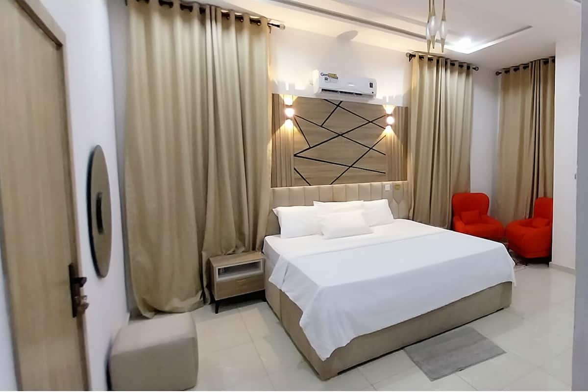 Habitación de hotel moderna con una cama grande, una mesita de noche, un pequeño mueble auxiliar, un aire acondicionado de pared y dos sillas rojas.