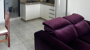 Eine kleine Küche mit weißen Schränken, einer Mikrowelle und einem Kühlschrank. Im Vordergrund steht ein violettes Sofa.