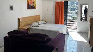 Ein Schlafzimmer mit Bett, Nachttisch, Kommode, einem Fenster mit Vorhängen und einem Balkon mit Blick.