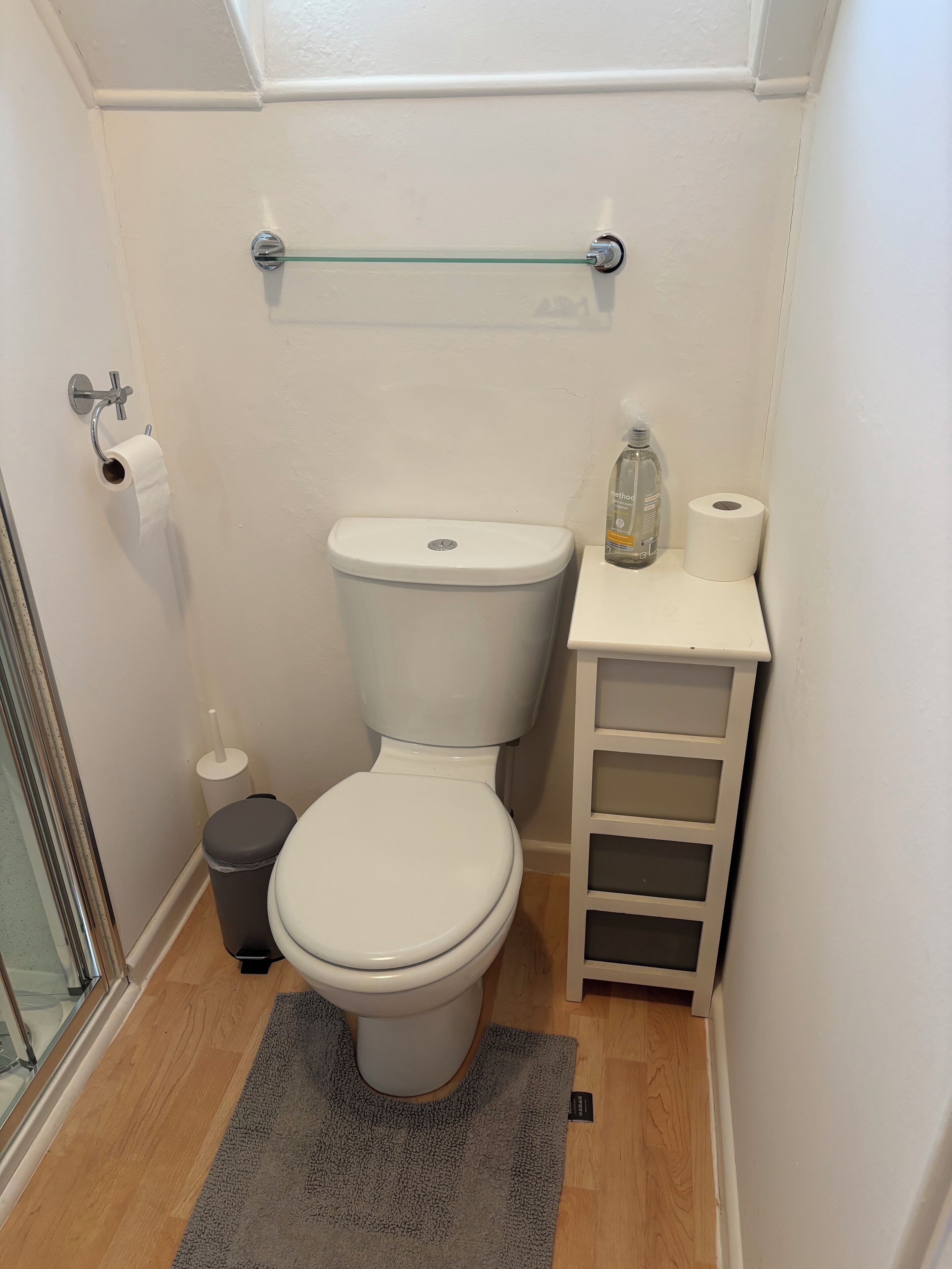 Ein kleines Badezimmer mit Toilette, einer Glasablage, einem Mülleimer und einem Toilettenpapierhalter.