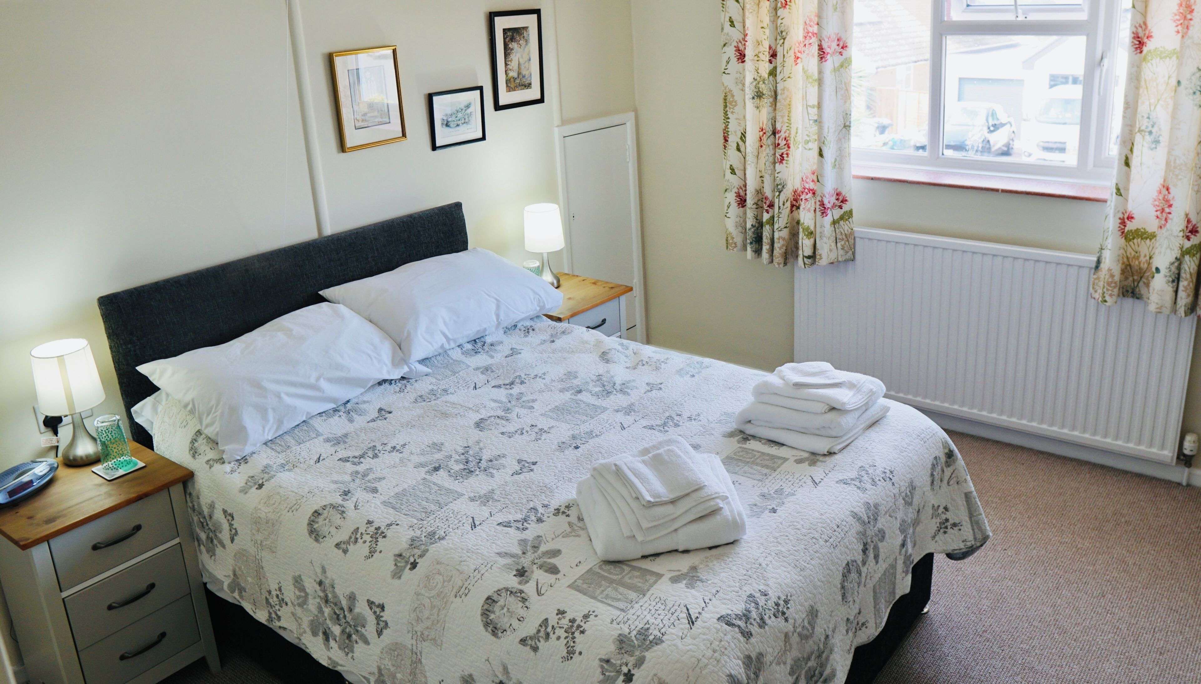A neatly made bed with a floral bedspread, a bedside lamp, a nightstand with a phone and a glass, and a window with floral curtains.