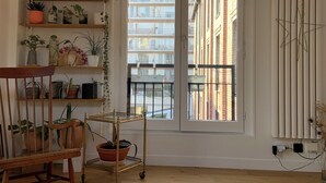 A room with wooden flooring, a chair, a wooden chair with a metal frame, a small cart, and a window with a view of buildings.