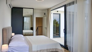 Une chambre d’hôtel moderne avec un grand lit, une table de chevet avec une lampe, un miroir, une porte-fenêtre coulissante donnant sur un balcon et une vue sur les arbres.