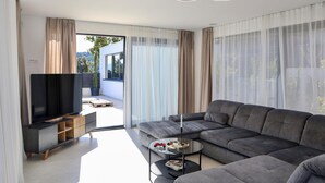 Un salon moderne avec un grand canapé d’angle gris, un téléviseur à écran plat posé sur un meuble en bois, et une table basse en verre sur laquelle se trouvent des télécommandes et une manette de jeu.