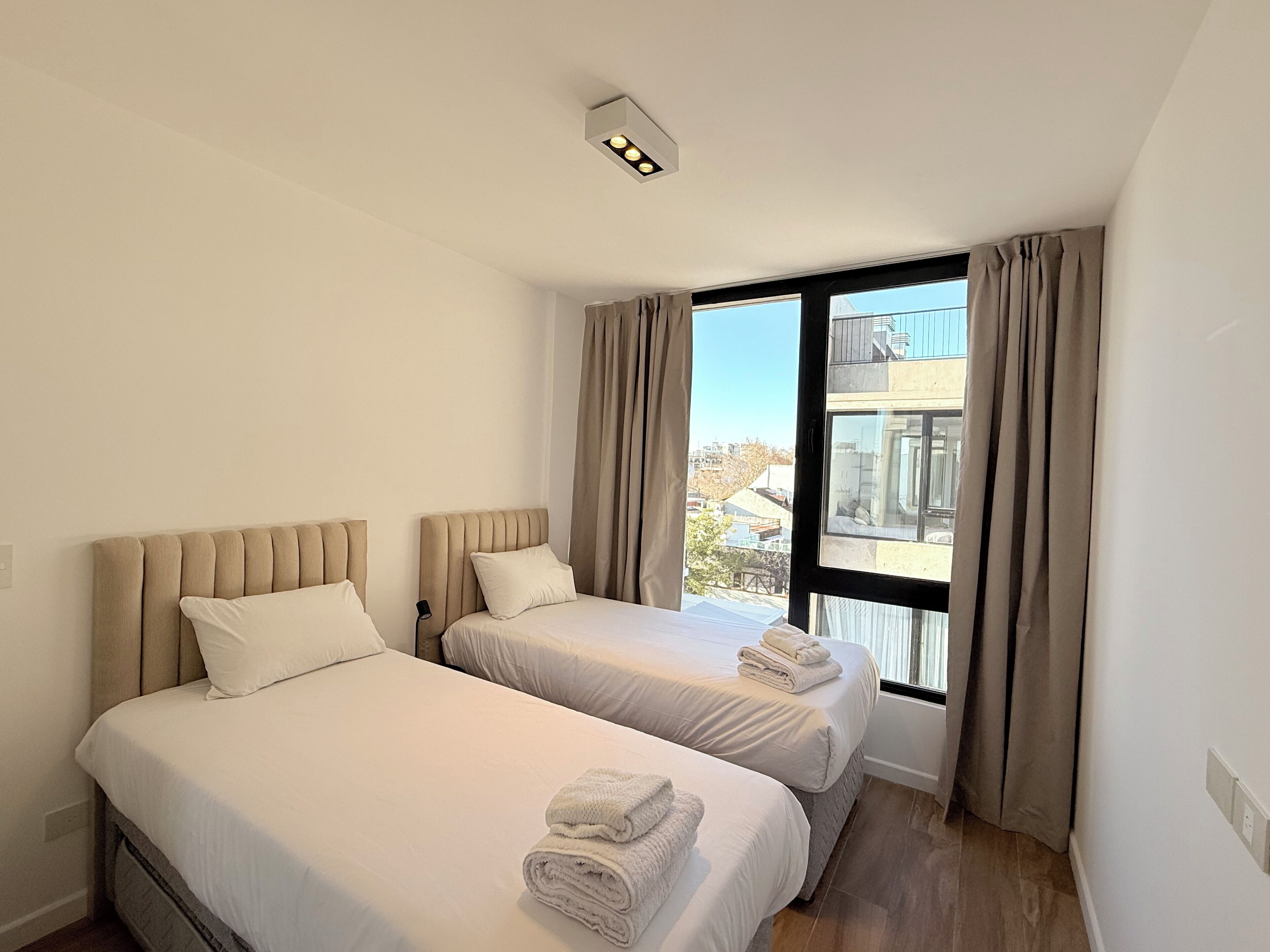 Habitación de hotel con dos camas, una ventana con cortinas y vistas a edificios.