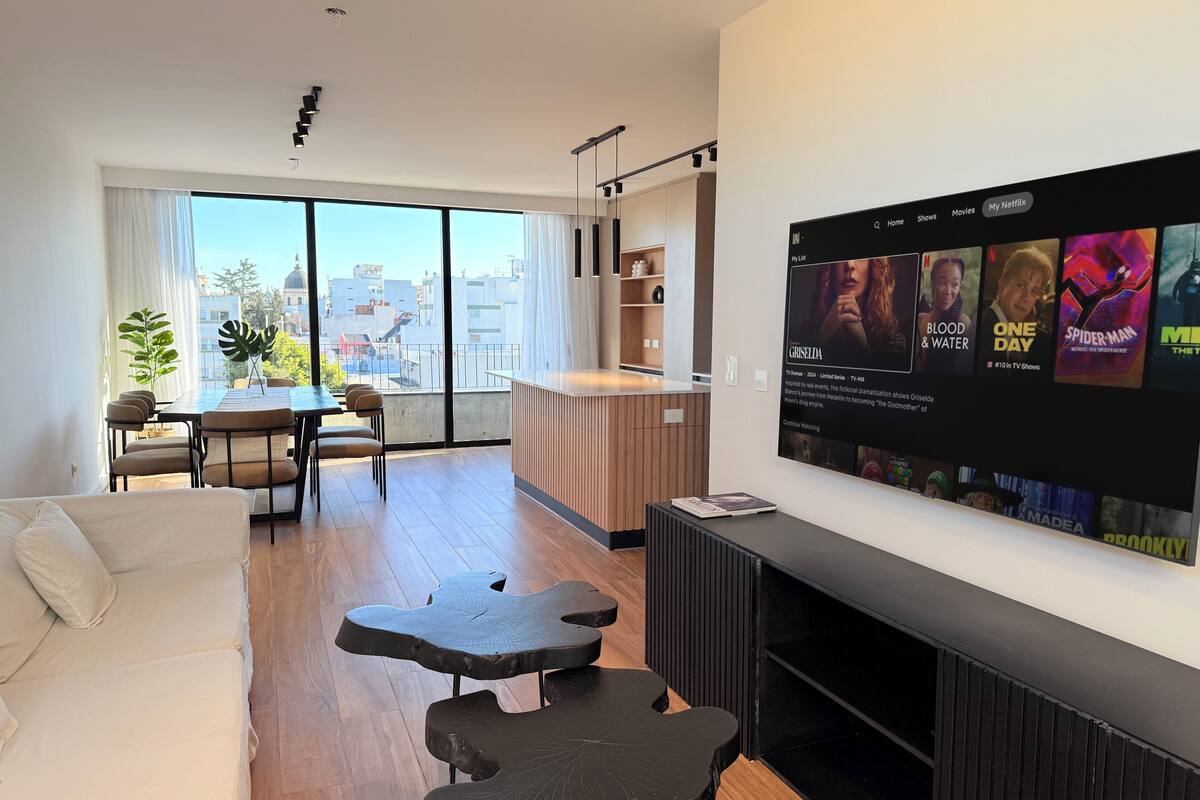Una sala de estar moderna con un televisor grande mostrando la interfaz de un servicio de streaming, un comedor con una mesa larga y sillas, y vistas de la ciudad a través de amplias ventanas.