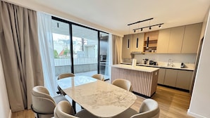 Una cocina moderna con una isla central, una zona de comedor con mesa de mármol y un amplio ventanal corredero que da a un balcón exterior.