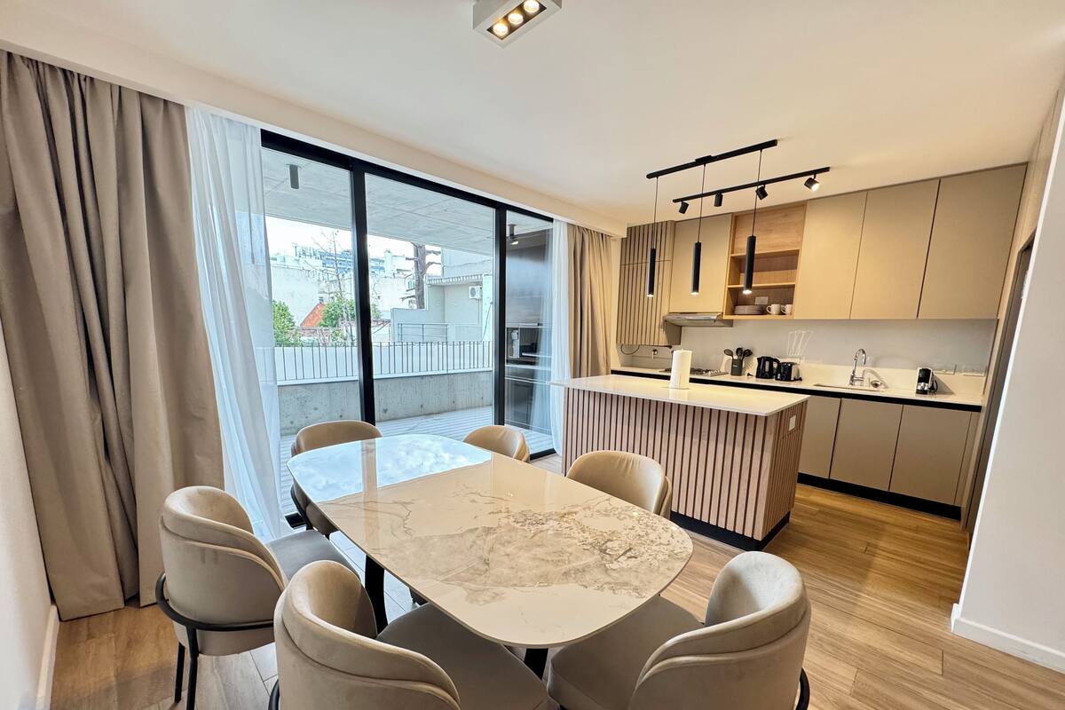 Una cocina moderna con una isla central, una zona de comedor con mesa de mármol y un amplio ventanal corredero que da a un balcón exterior.