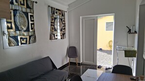 A modern living room with a grey sofa, a wooden dining table, and a chair. There is a door leading outside, a small table with a bottle, and a wall-mounted shelf. - Golden Papaya  (Sainte-Rose)