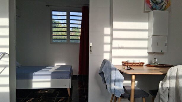 A compact room with a bed, a small table with a chair, a window with blinds, and a wall-mounted printer. - Golden Papaya  (Sainte-Rose)