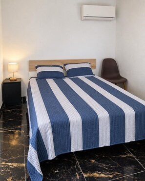 A hotel room with a bed, a nightstand, a lamp, a chair, and an air conditioning unit.